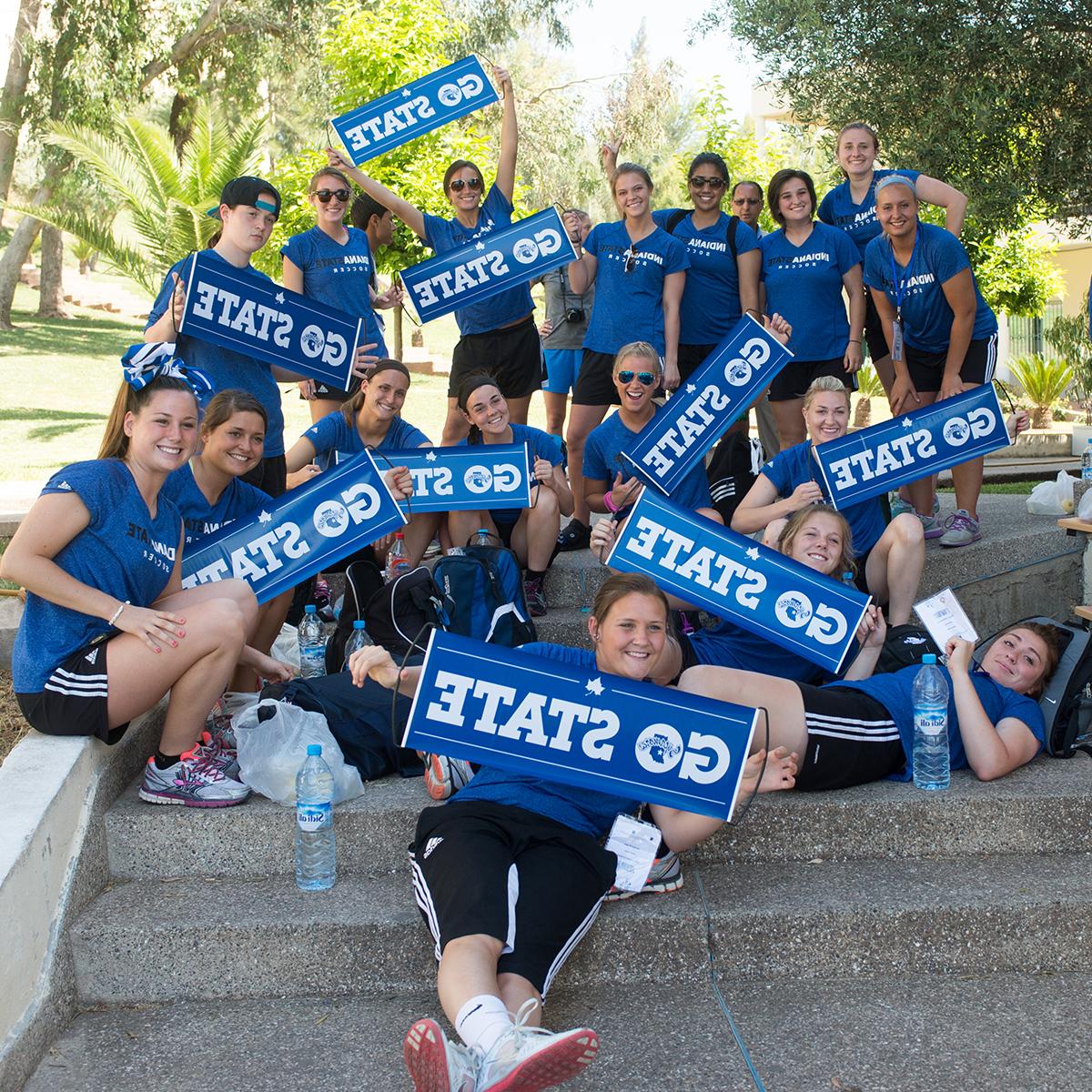 A group of students and staff clad in blue shirts and black pants hold up blue signs with mantras such as "God blue!" printed on them. They are sitting near one another in a stairway on a small hill with tropical vegetation swaying gently in the wind in the background.